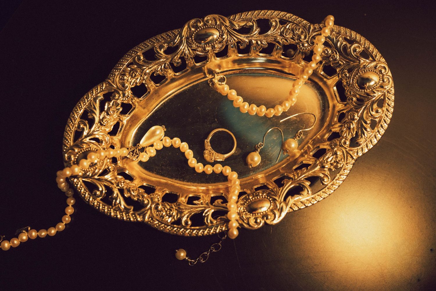 Ornate gold filigree oval tray with pearl necklace and ring displayed on mirrored surface with warm lighting.
