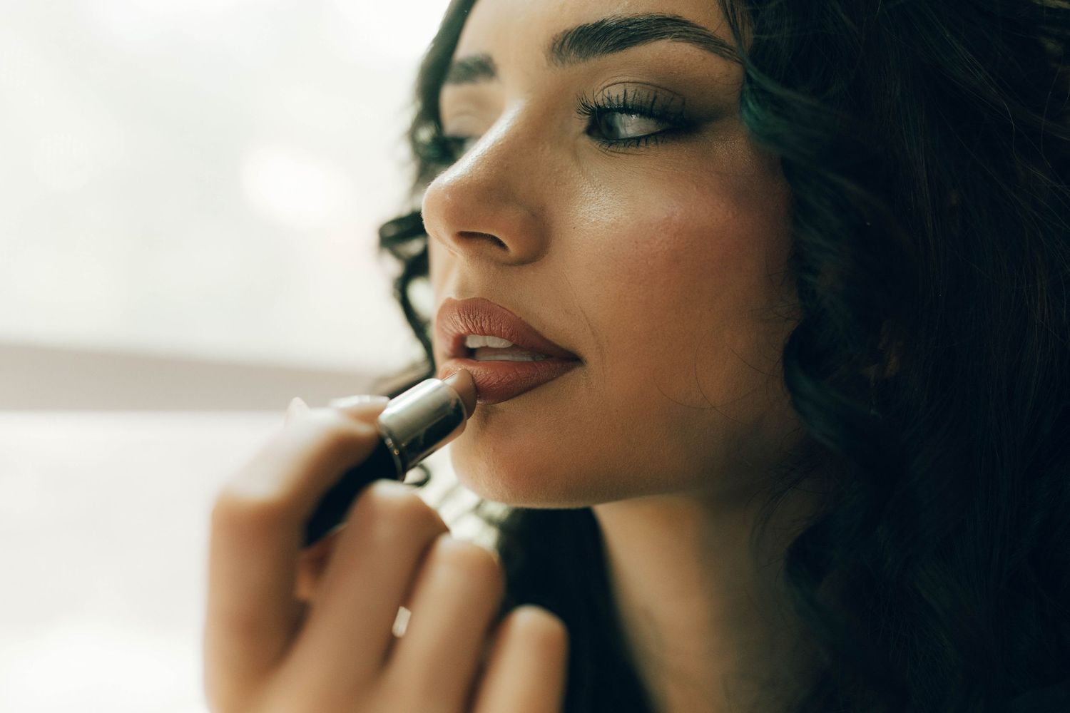 Young woman applying nude lipstick with a sleek black tube, showcasing elegant makeup artistry.