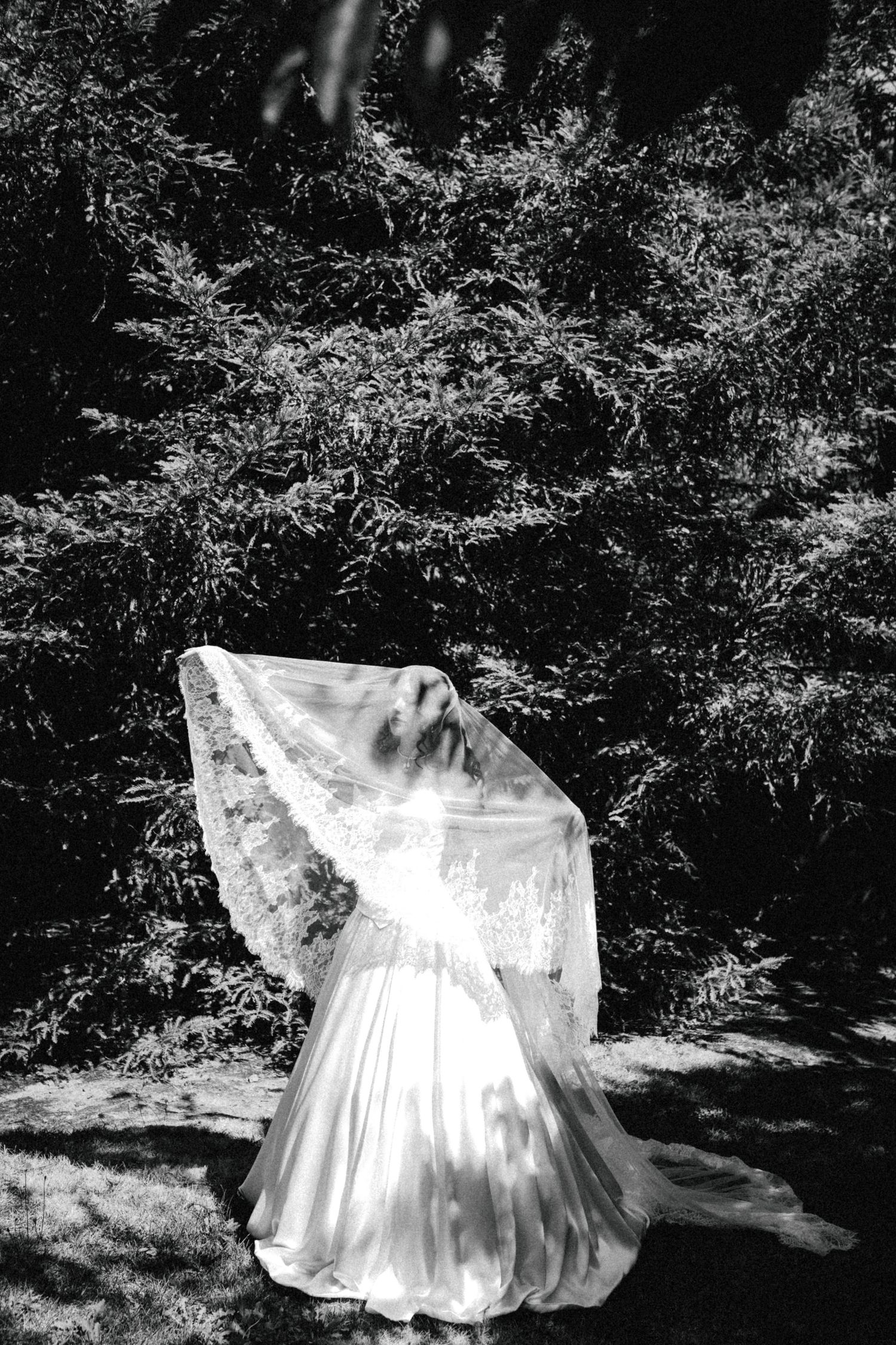 Black and white photo of a bride in a flowing gown and lace veil outdoors among lush green trees.