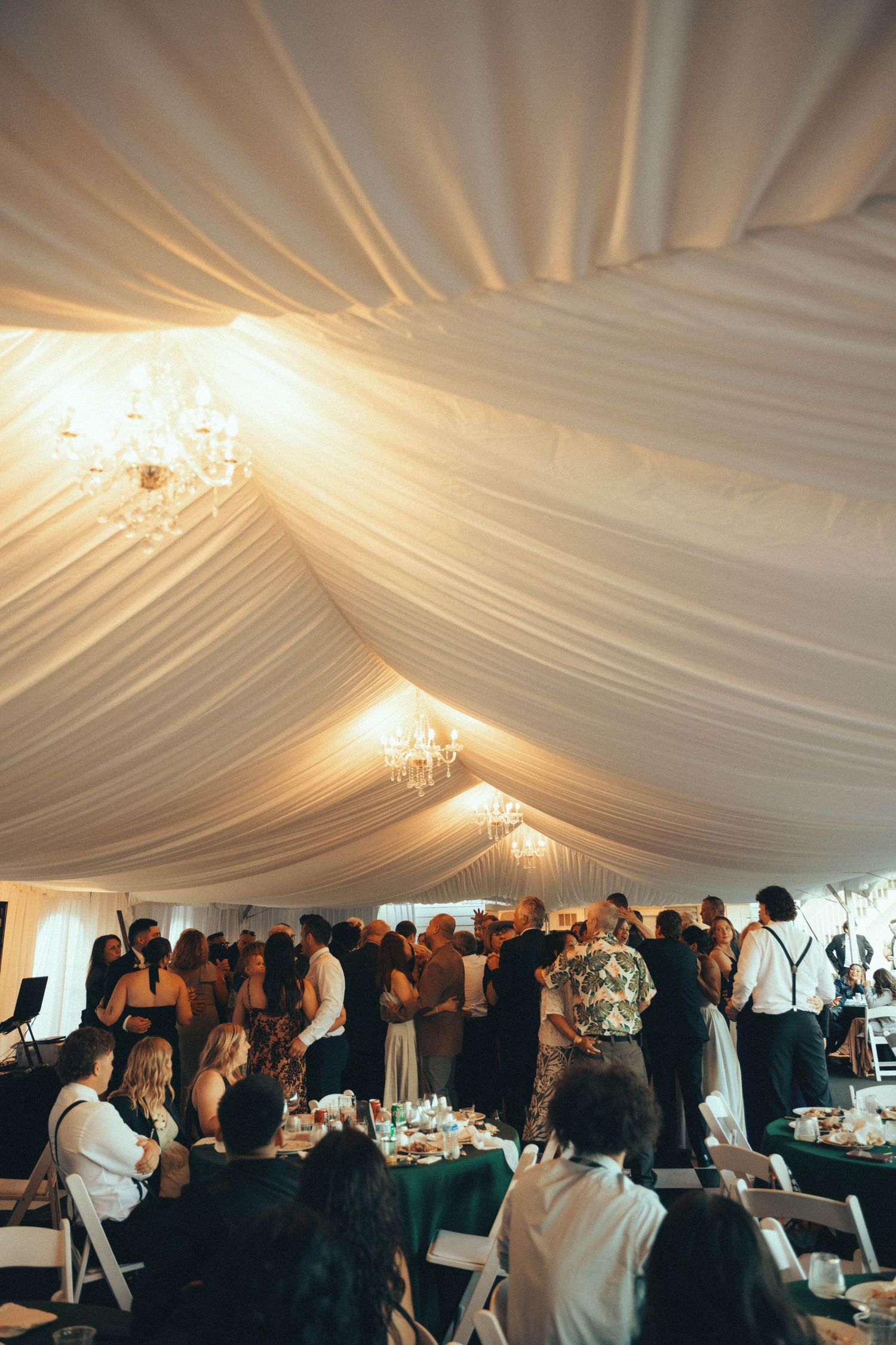 Elegant wedding reception under a white draped tent with chandeliers, guests mingling and seated at green-accented tables.