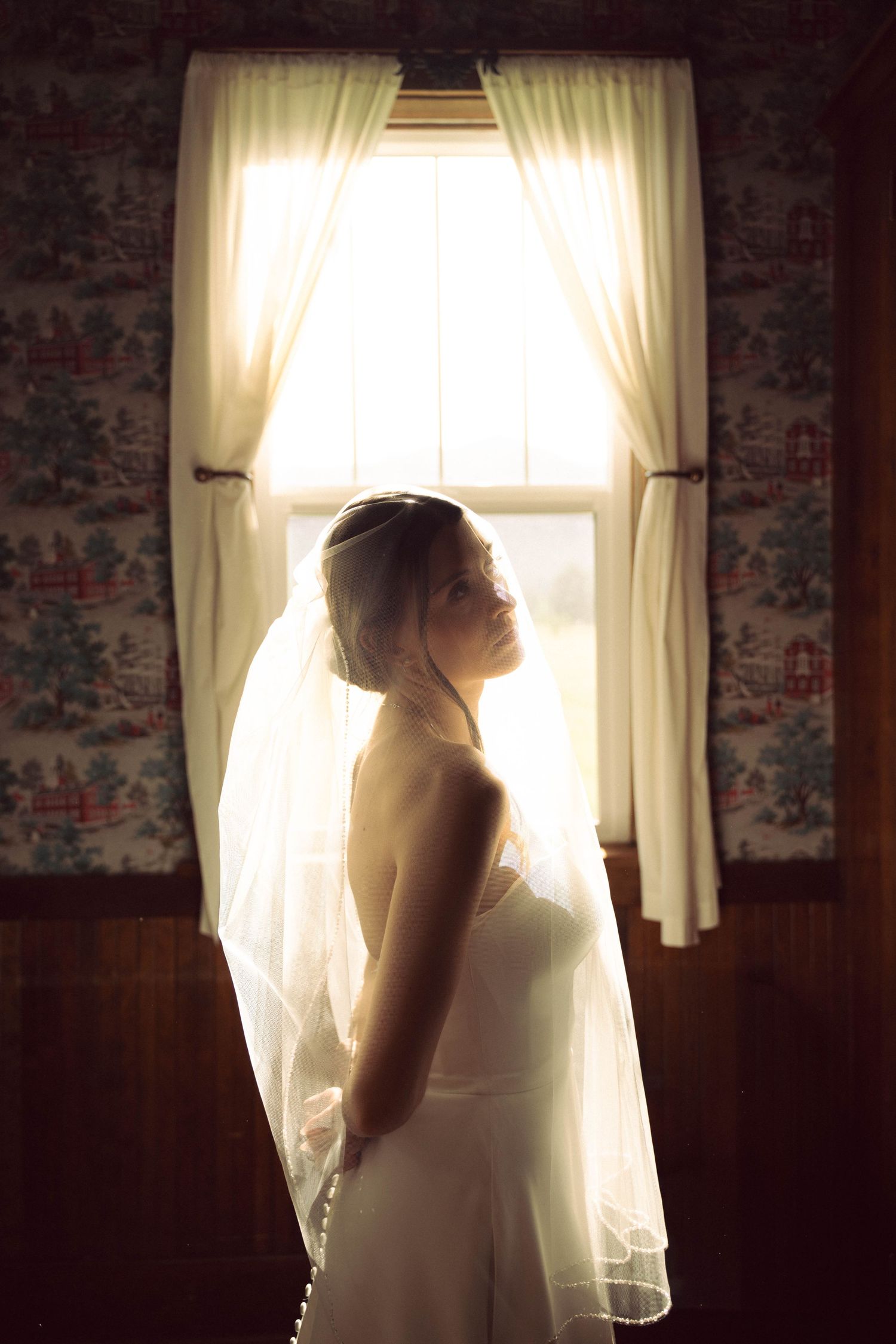 Bride in white gown and veil stands silhouetted by bright window light in a warmly lit room.