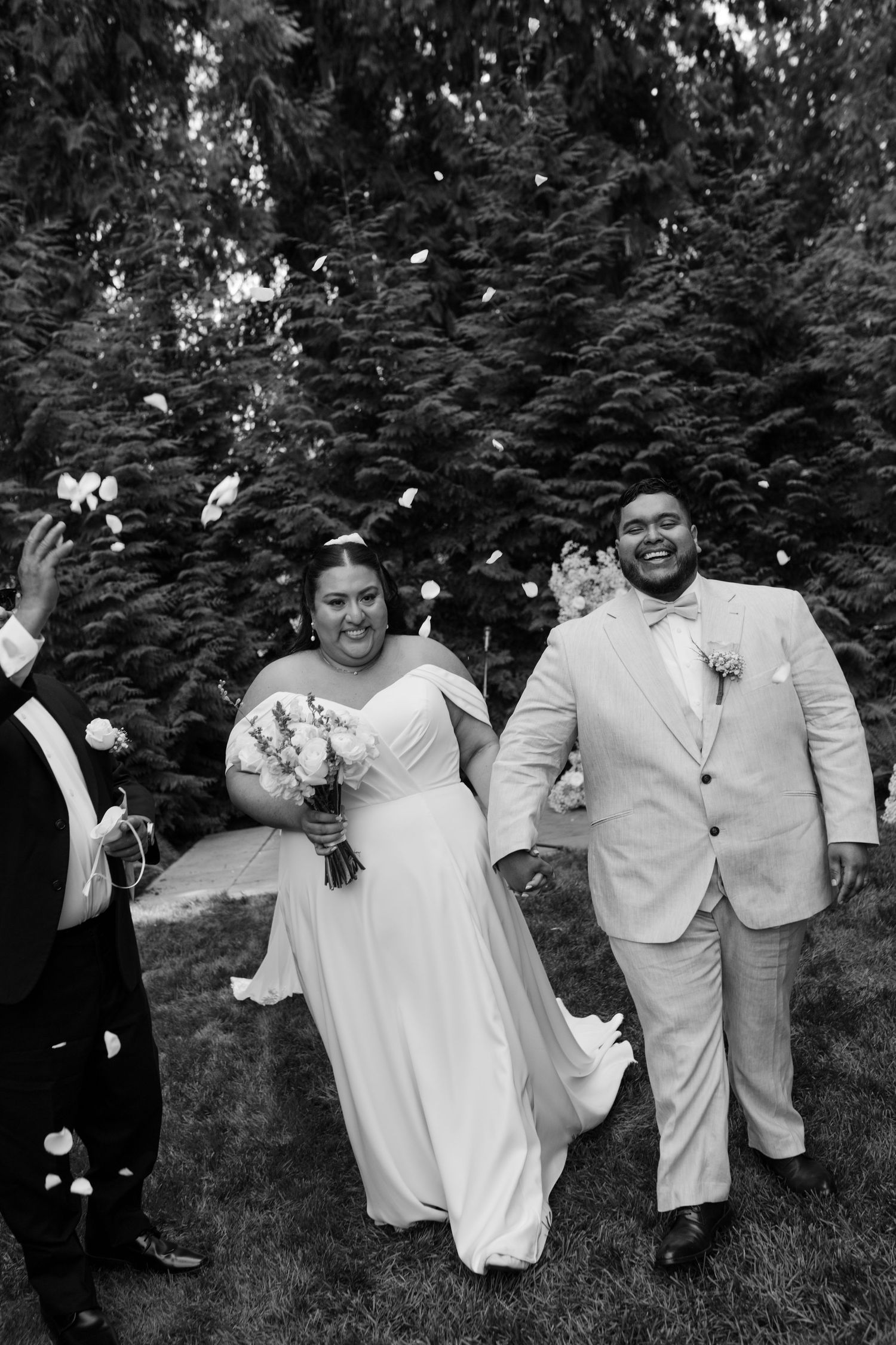 Three joyful people in white attire laugh and embrace while celebrating outdoors in a garden setting.