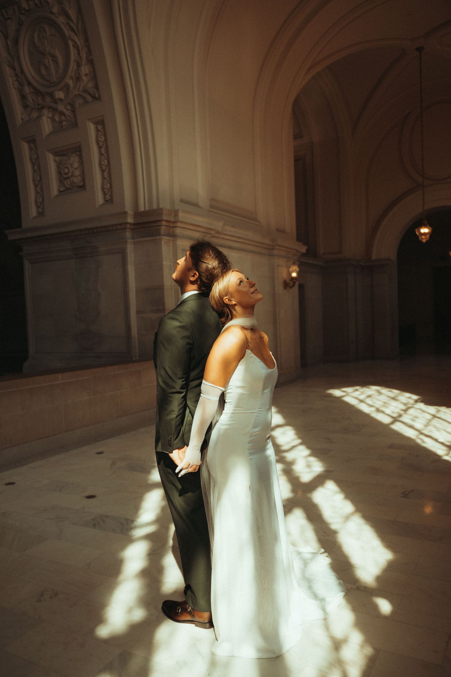 A couple shares a romantic kiss in an elegant marble hallway as sunlight streams through windows creating dramatic shadows.
