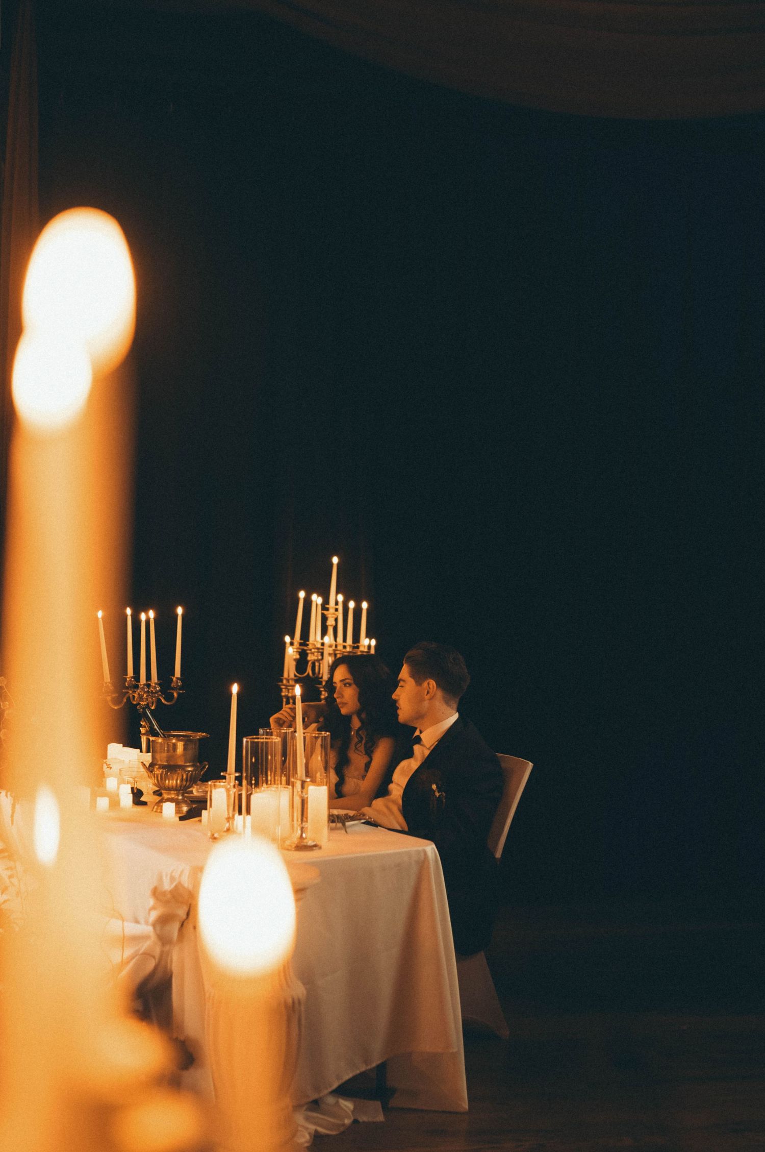 Couple seated at candlelit sweetheart table with elegant candelabras during romantic wedding reception dinner.