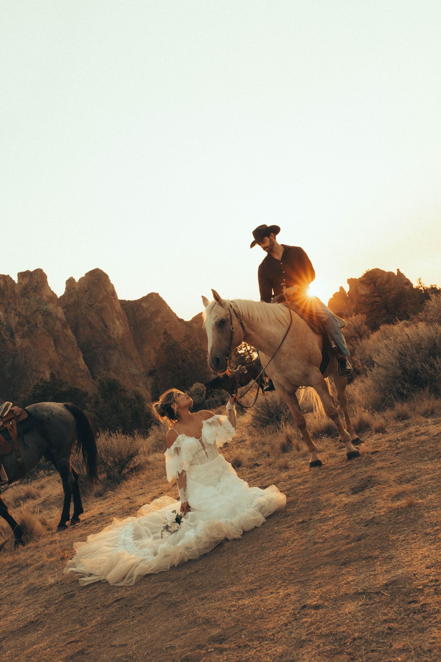 Bride in white gown sits on desert hillside as groom on white horse reaches down at golden sunset.