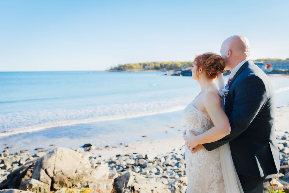 Newlyweds embrace on a scenic rocky beach with blue ocean waves in the background.