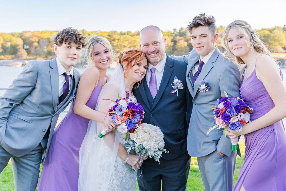 Wedding party poses together outdoors wearing purple bridesmaid dresses and gray suits.