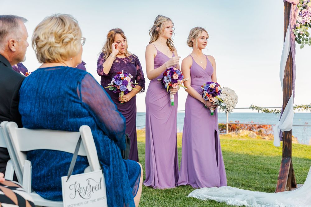 Wedding ceremony by the water with bridesmaids in purple dresses watching the proceedings.