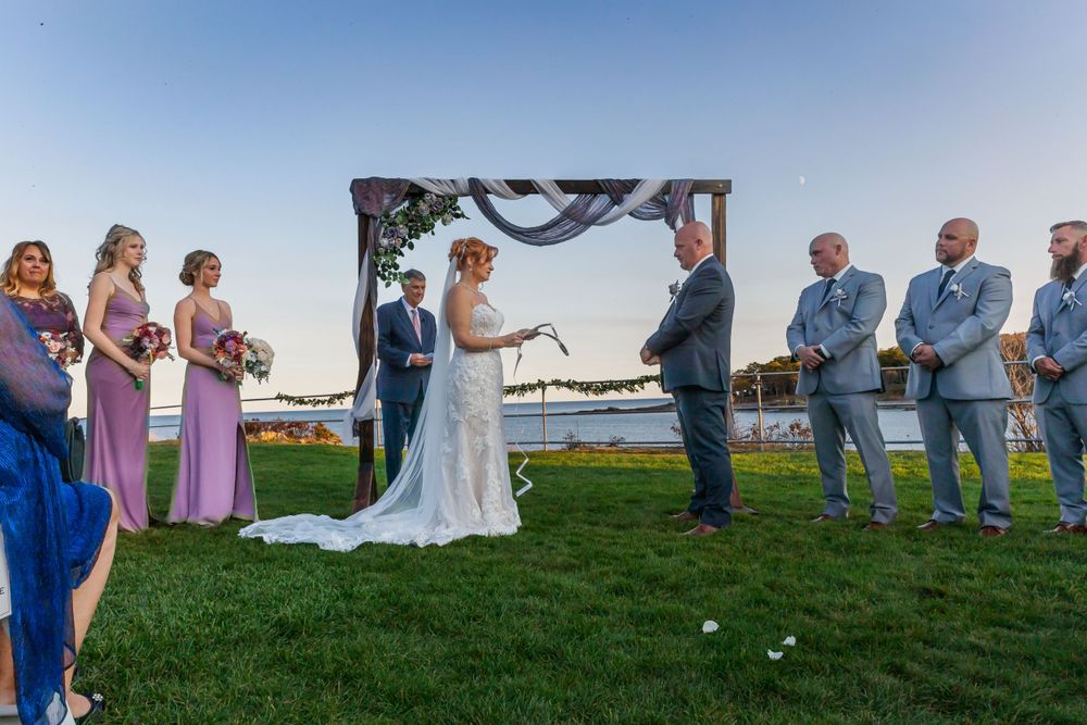 Outdoor waterfront wedding ceremony under decorated arch with bridal party and guests.