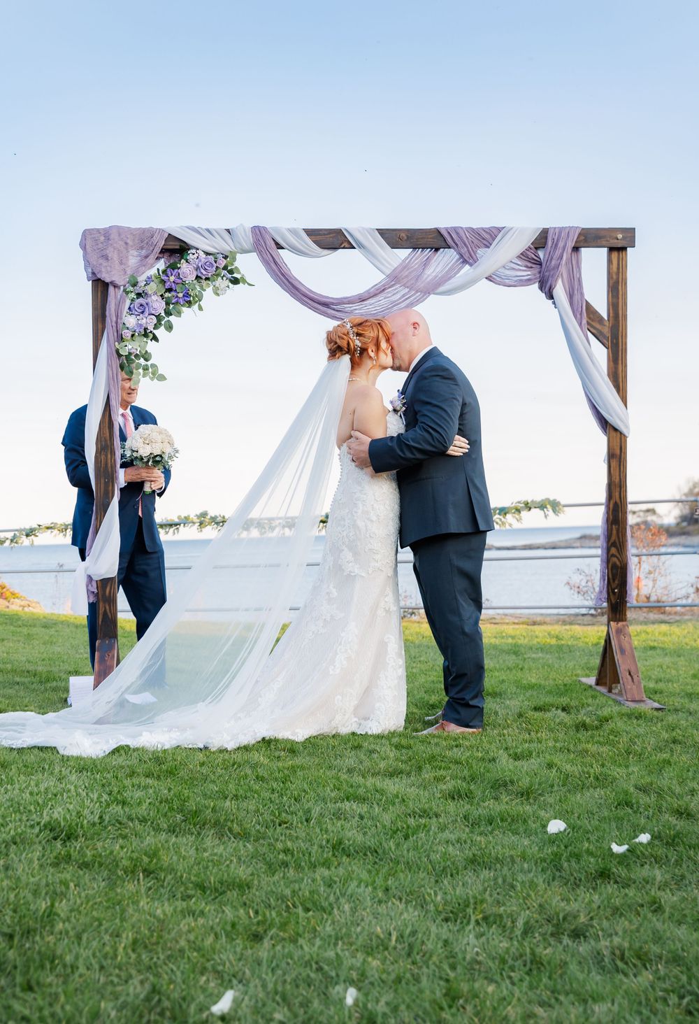Wedding ceremony kiss under a decorated wooden arch with waterfront views.