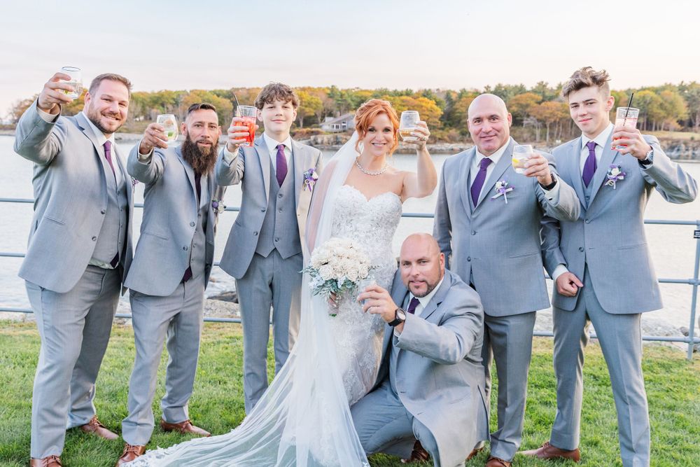 Fun wedding party photo with groomsmen in light gray suits posing by the water.
