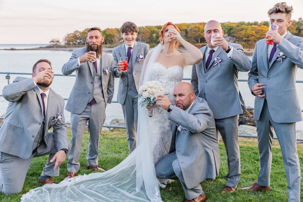 Playful wedding party strikes humorous poses on grass near waterfront.