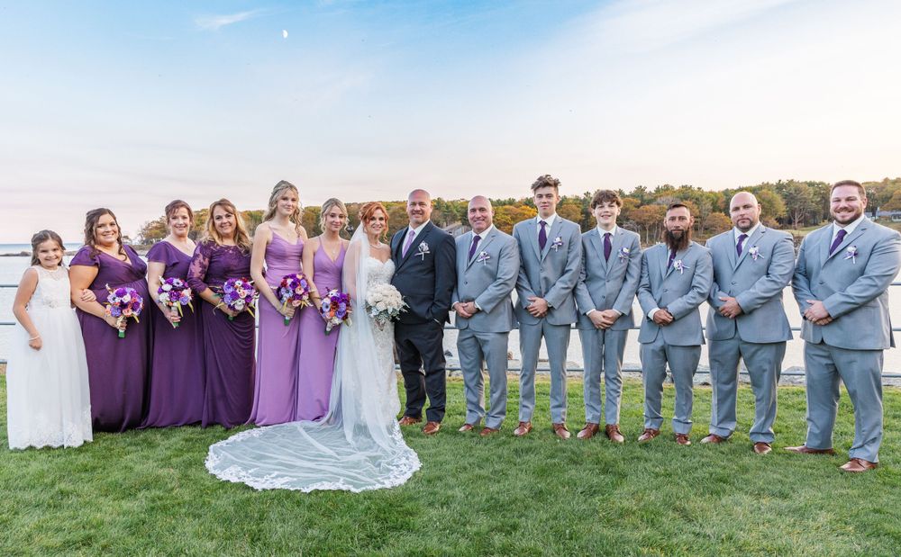 Bridal party posed on grass with bridesmaids in purple and groomsmen in gray suits by water.
