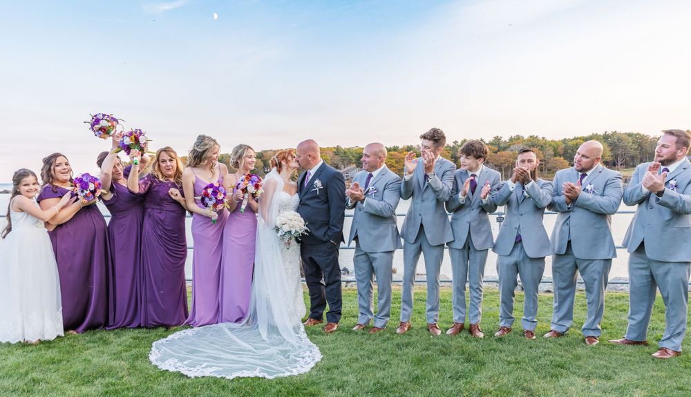 Wedding party lined up outdoors with bridesmaids in purple dresses and groomsmen in light gray suits.