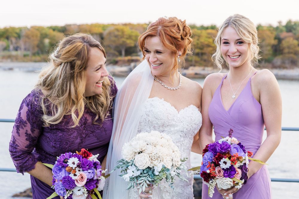 Three women wearing purple and lavender dresses hold bouquets with purple and white flowers.