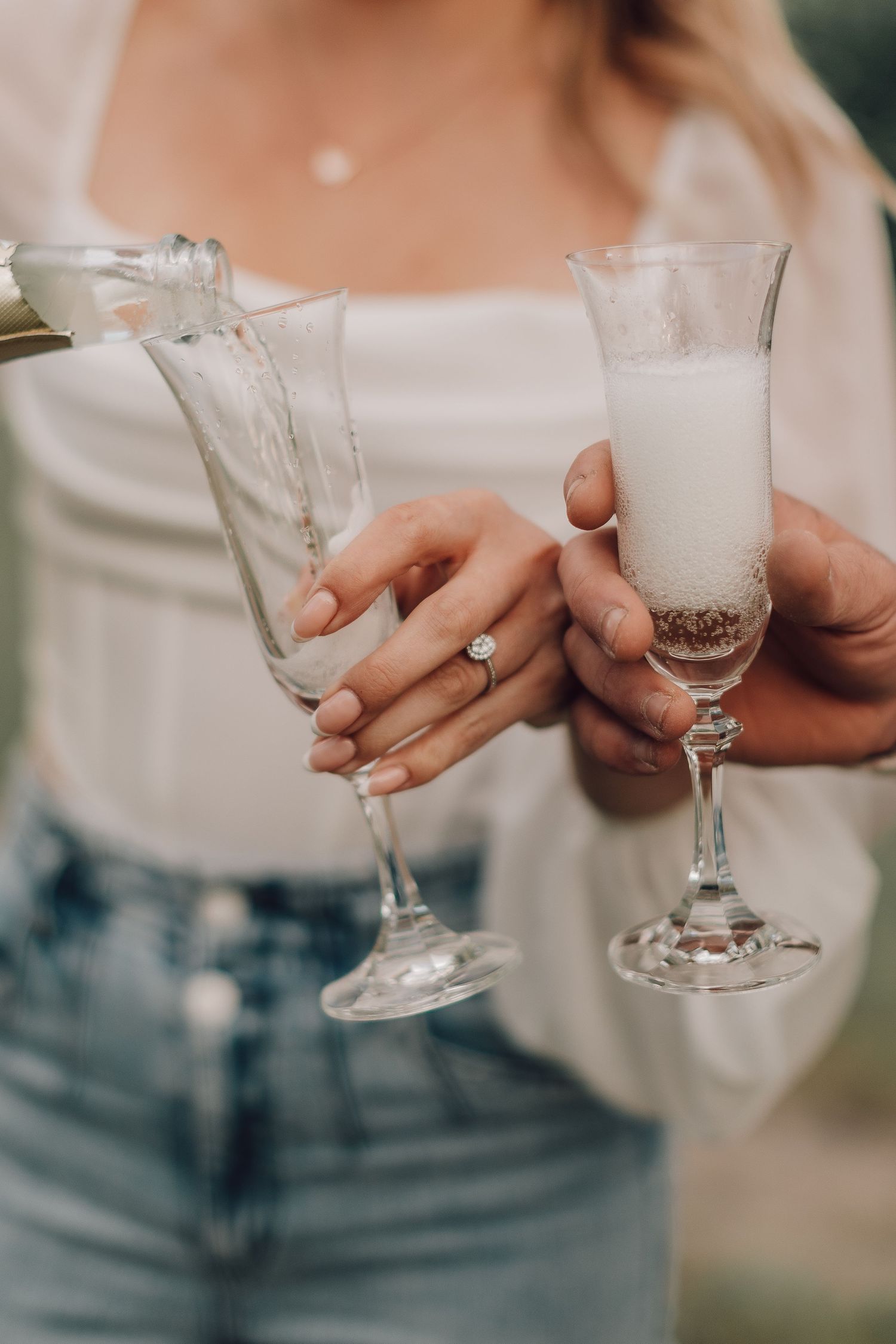 Close-up of engaged couple pouring champagne into crystal flutes outdoors.