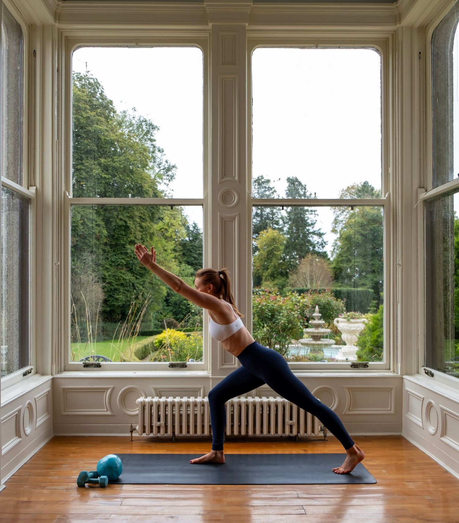 Yoga practitioner performing warrior pose in elegant room with large bay windows.