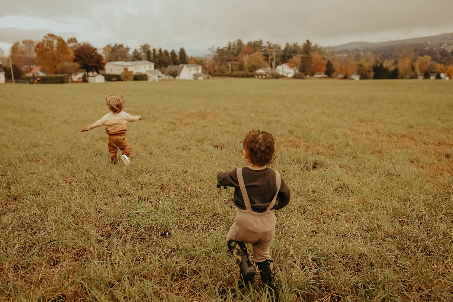 Two children running and playing in an open autumn field with farmland in the background.