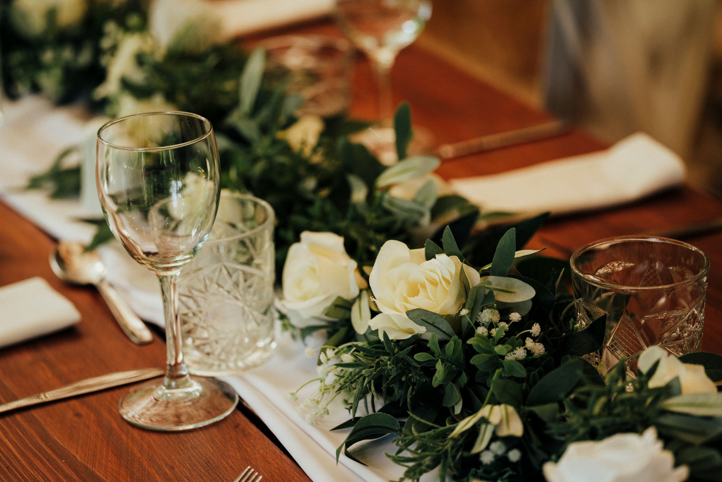 Wedding table setting with white roses, greenery garland and crystal glassware on wooden table.