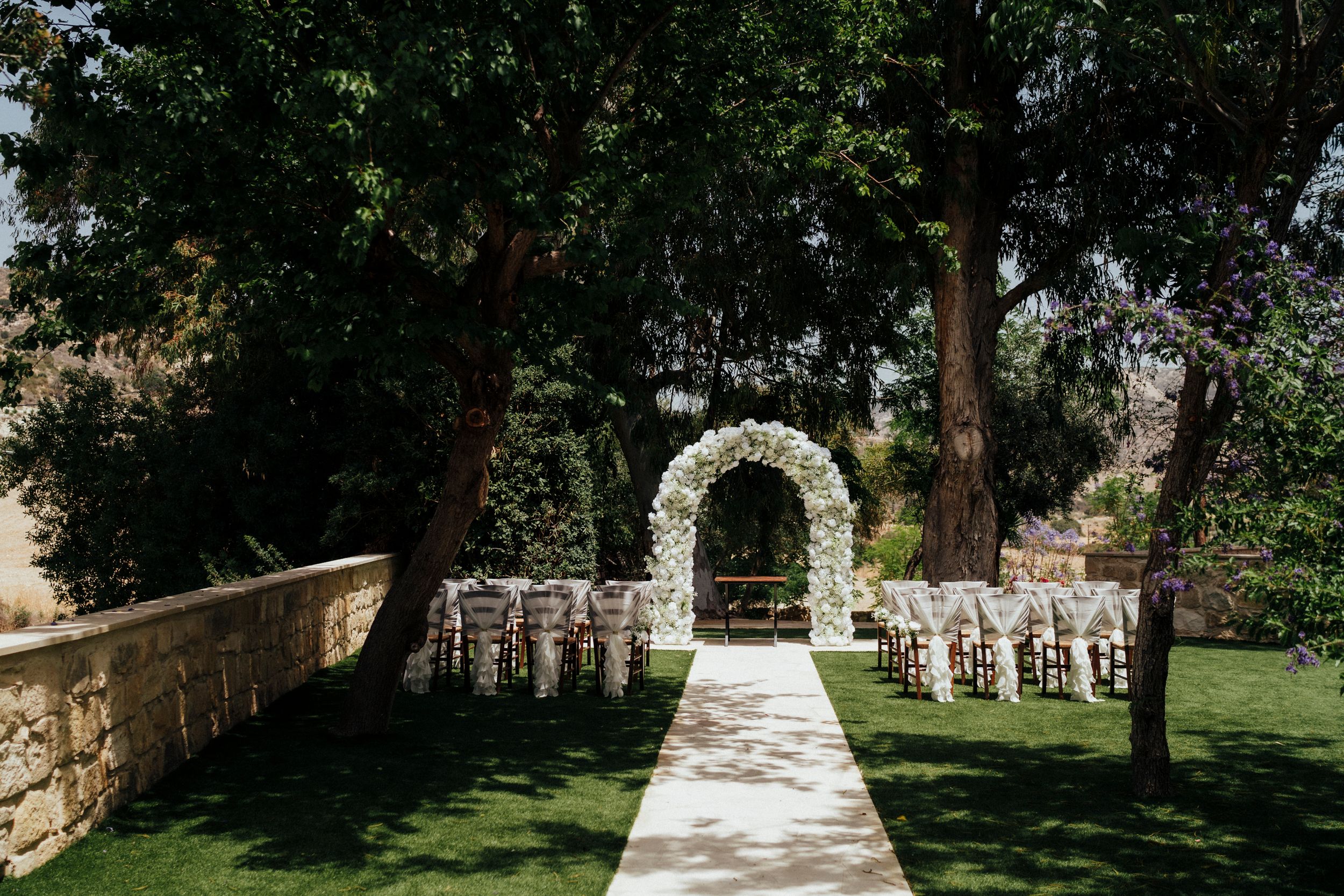 Garden wedding ceremony site with white floral arch and rows of chairs on manicured lawn.