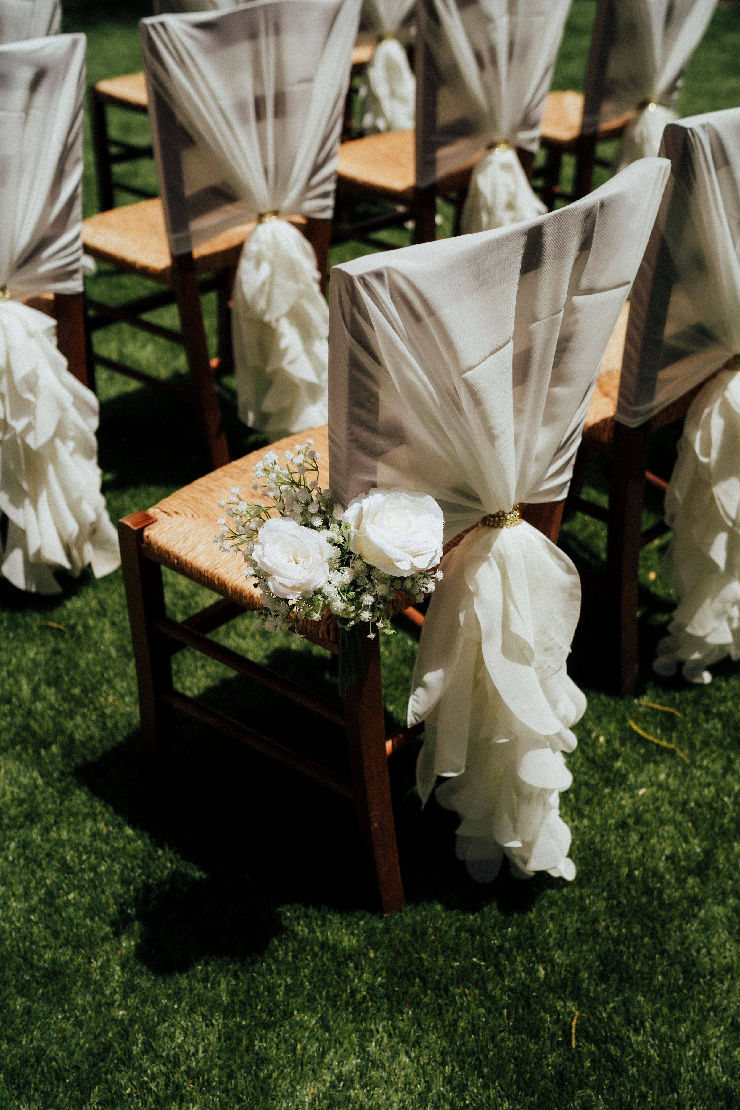 White chair covers with floral decorations arranged on wooden chairs for an outdoor wedding.