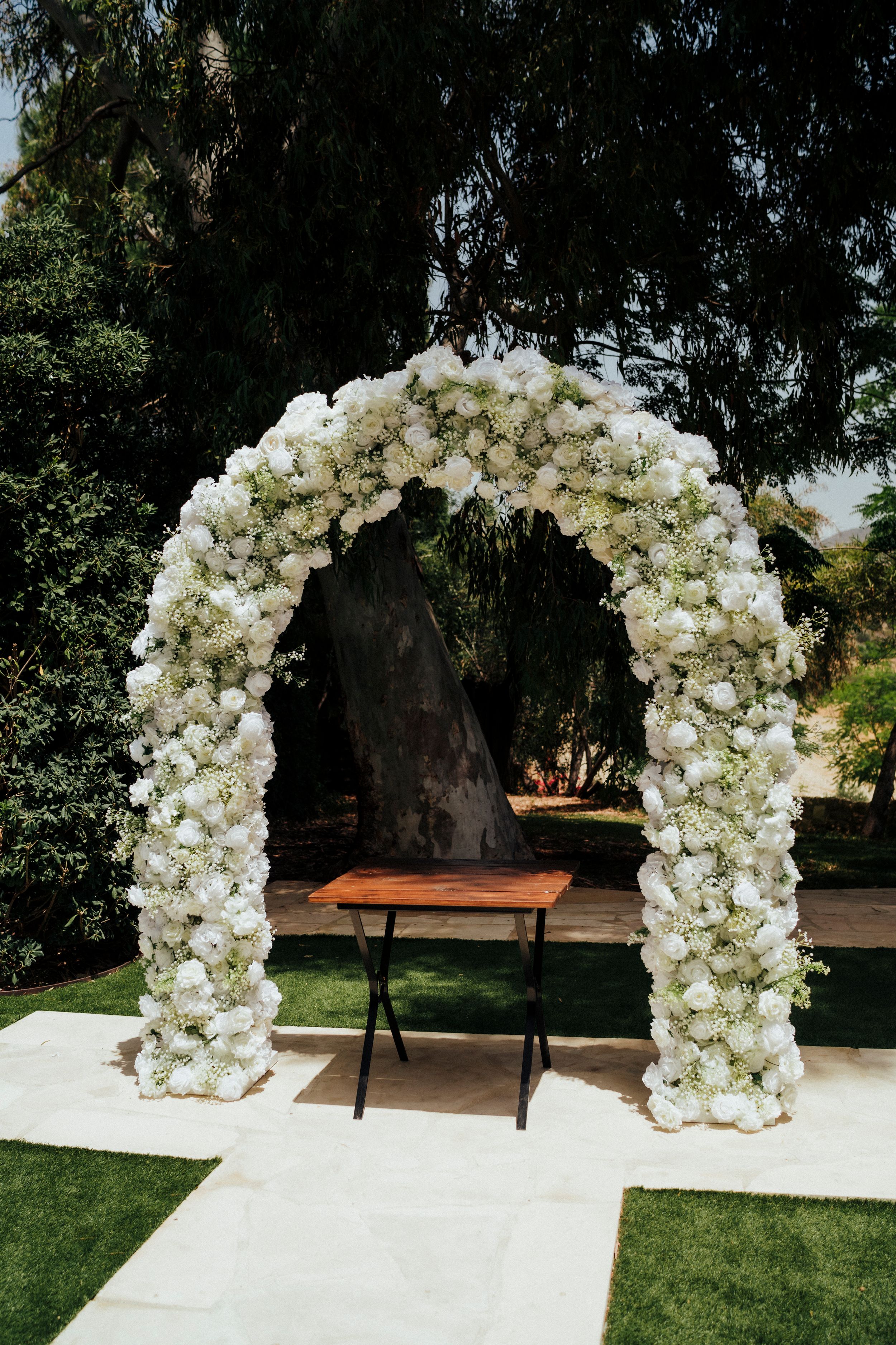 Stunning white rose arch with dense floral arrangement stands on stone pathway in garden setting.