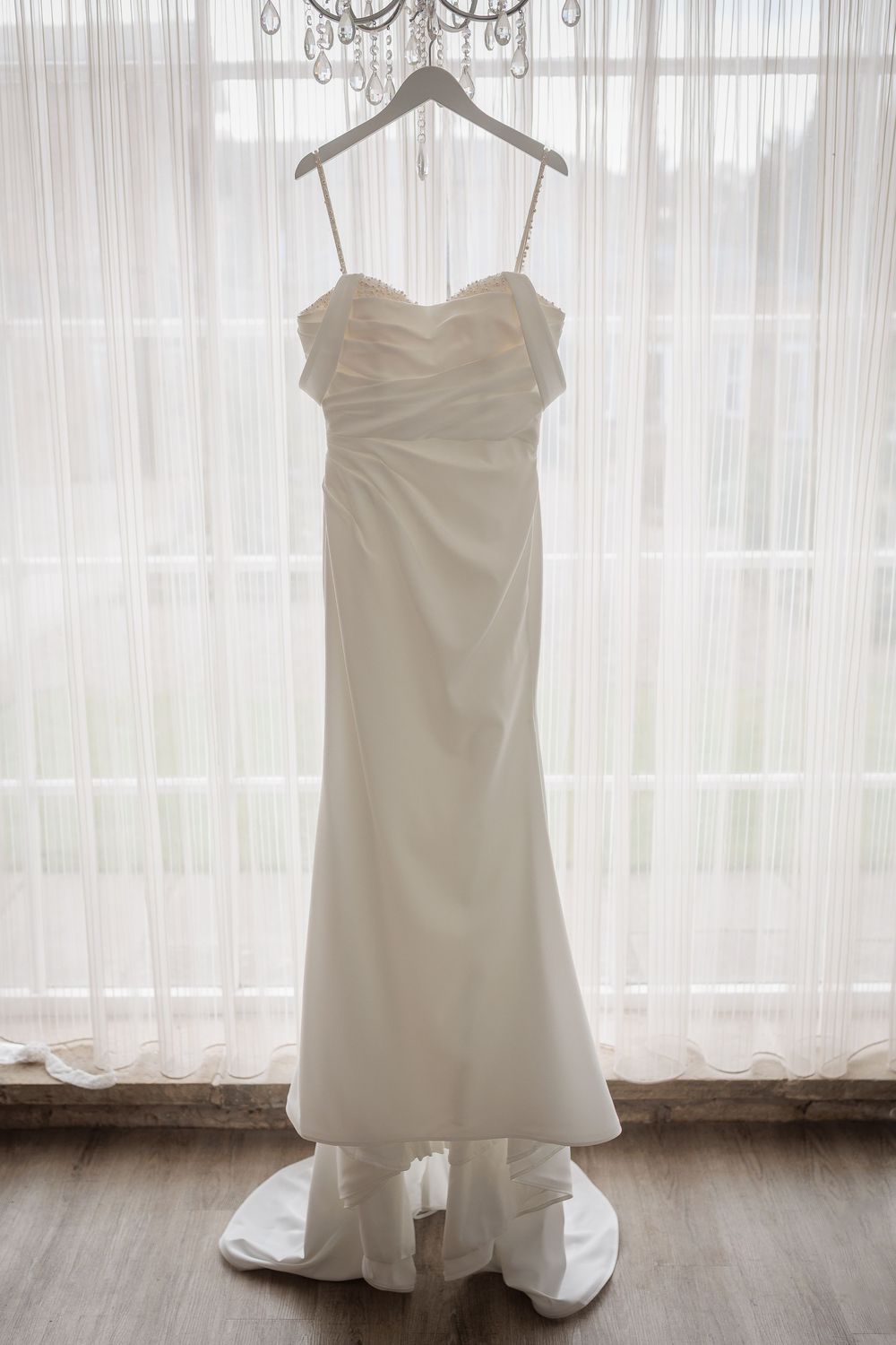 A white wedding dress hangs elegantly in front of a sunlit window with sheer curtains creating a romantic silhouette.