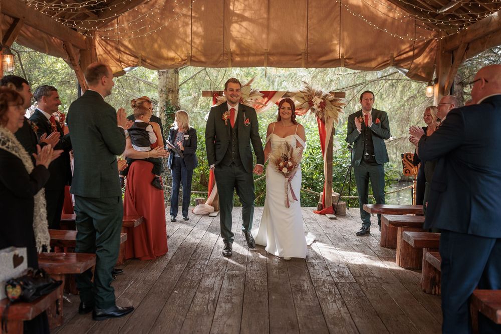 A panoramic view of a rustic wooden covered bridge decorated for a wedding ceremony with guests gathering inside.