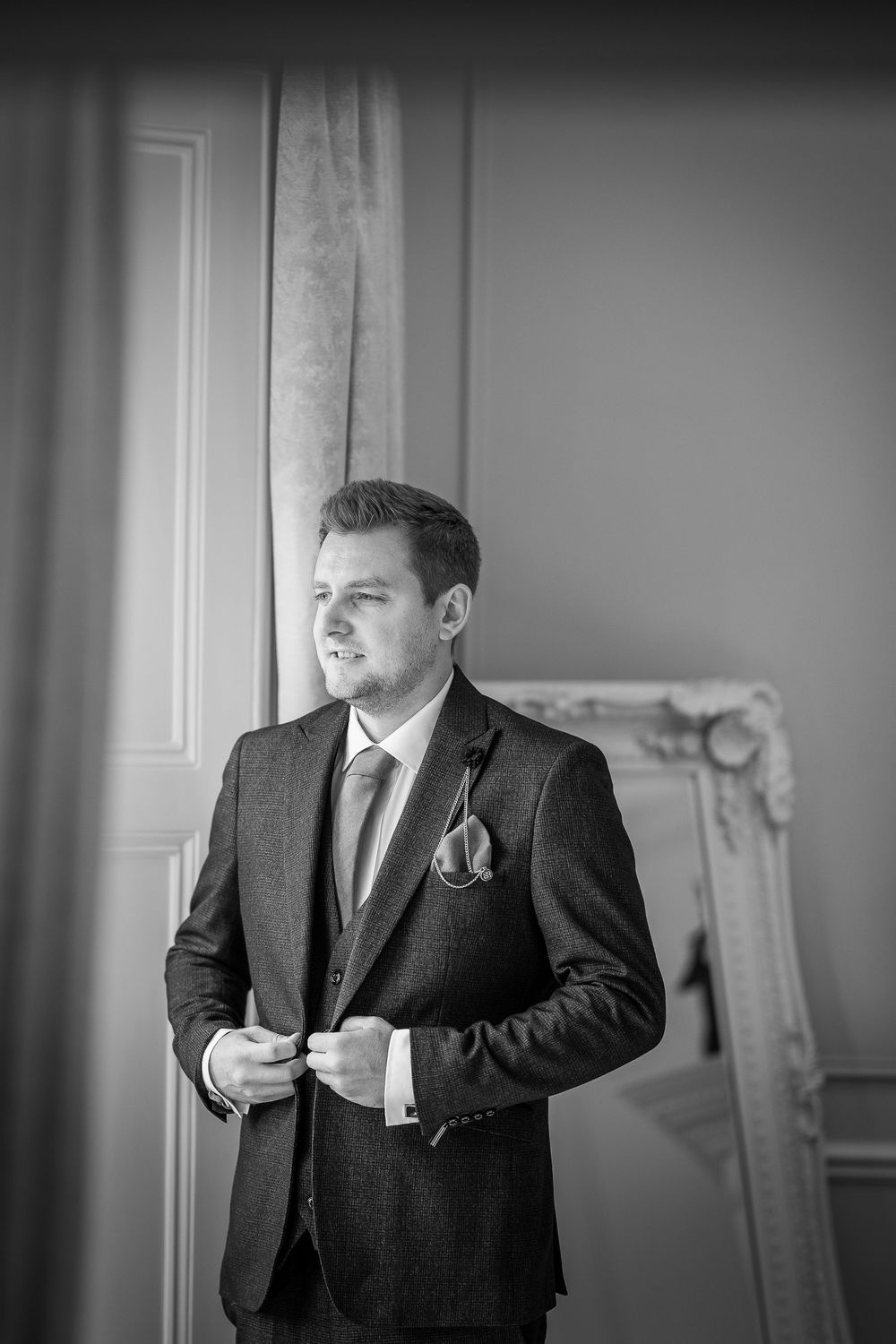 Black and white portrait of a groom in a classic dark suit adjusting his tie while standing by a window.