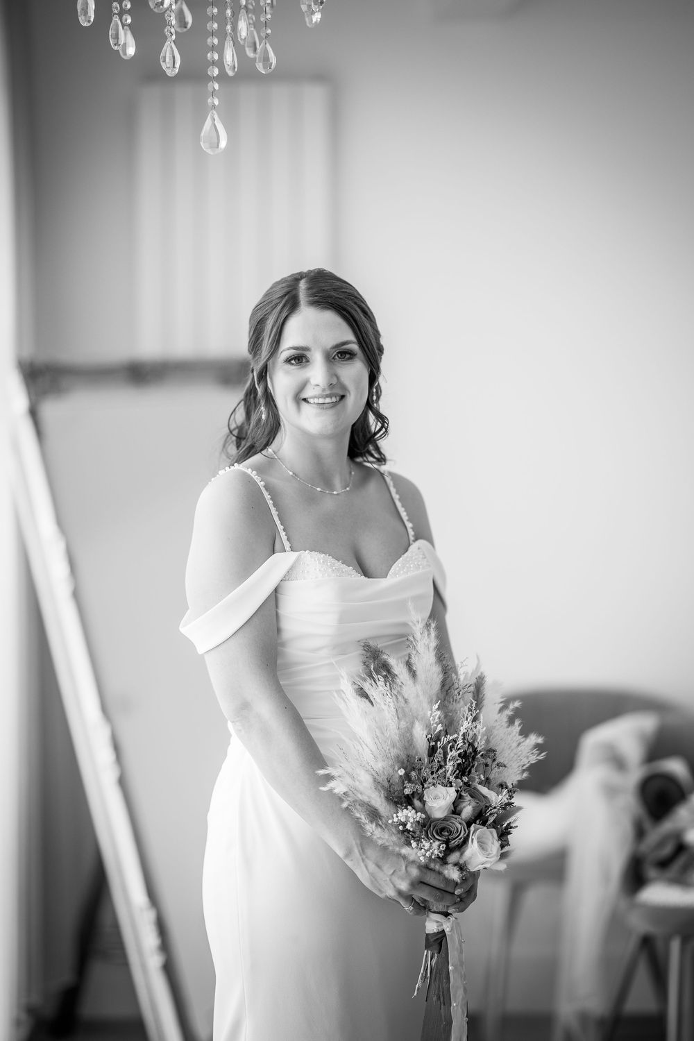 A black and white wedding photo showing a bride in an elegant off-shoulder dress holding a dried flower bouquet.