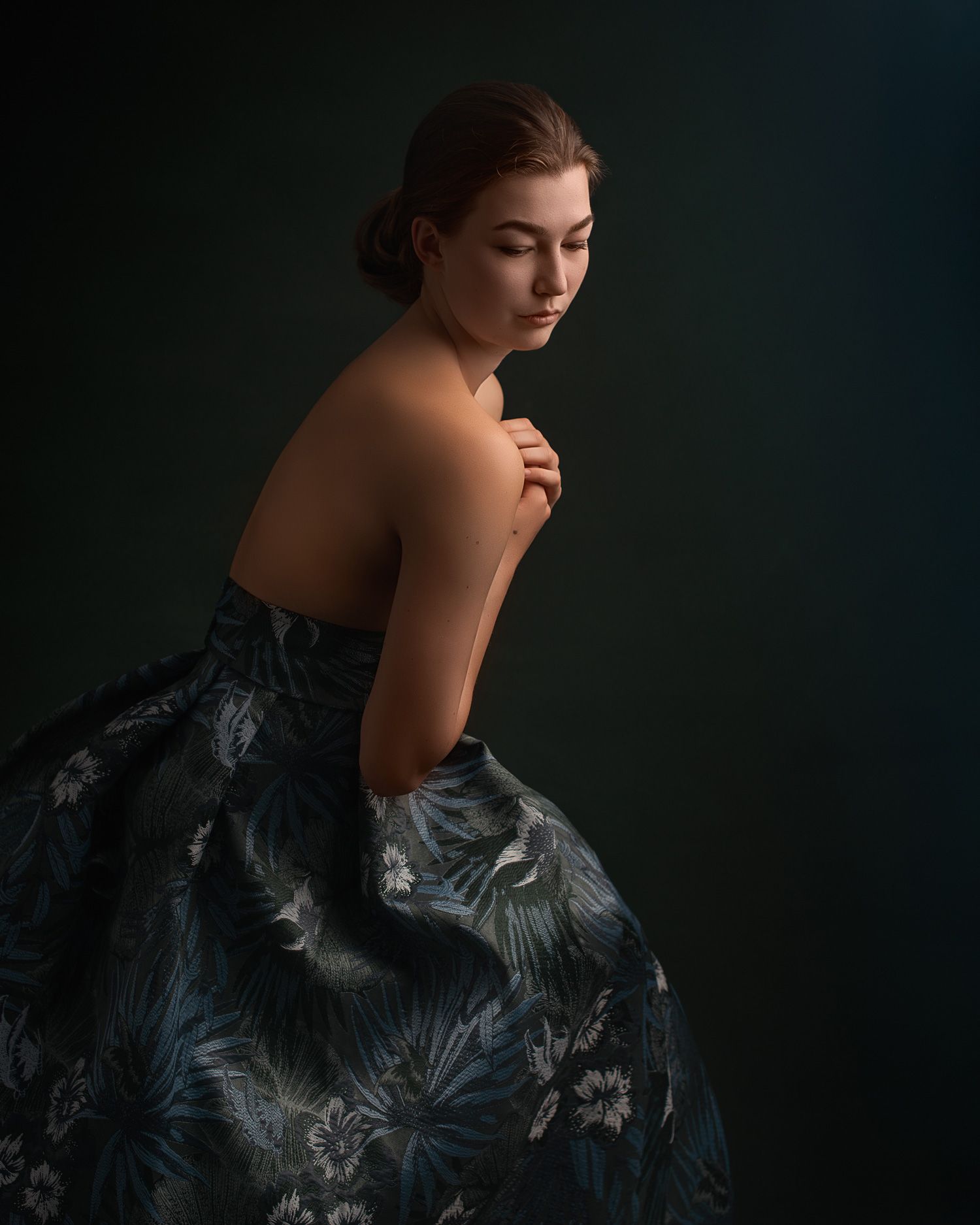 Moody portrait of someone in an elegant floral evening gown against a dark background with dramatic lighting.