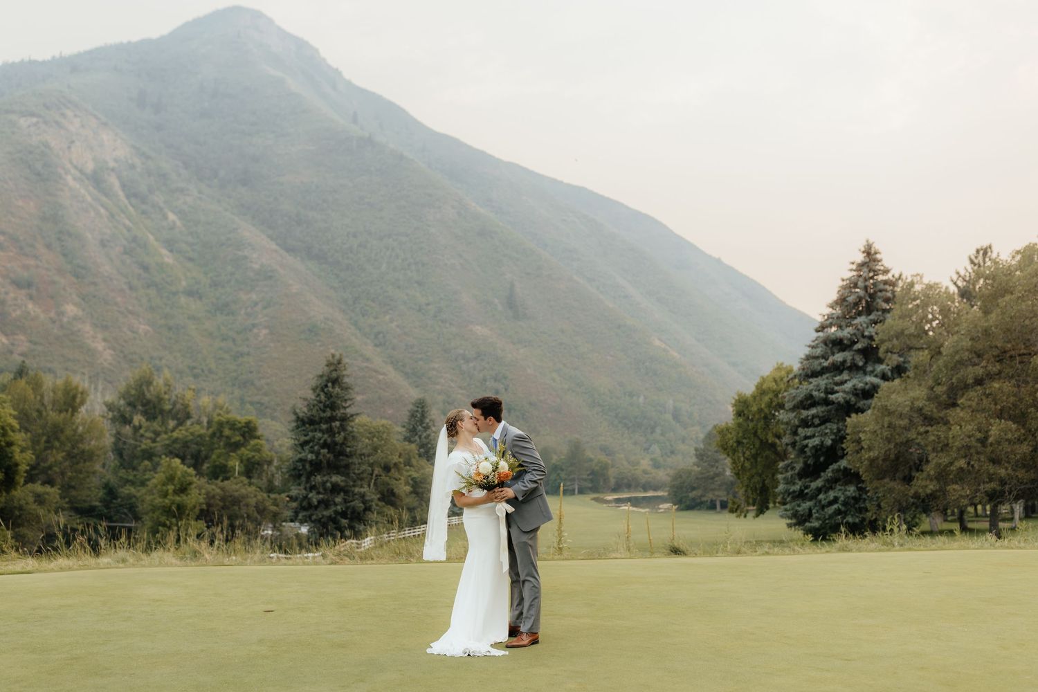 Bride and groom share a romantic kiss on a green golf course with a majestic mountain backdrop.