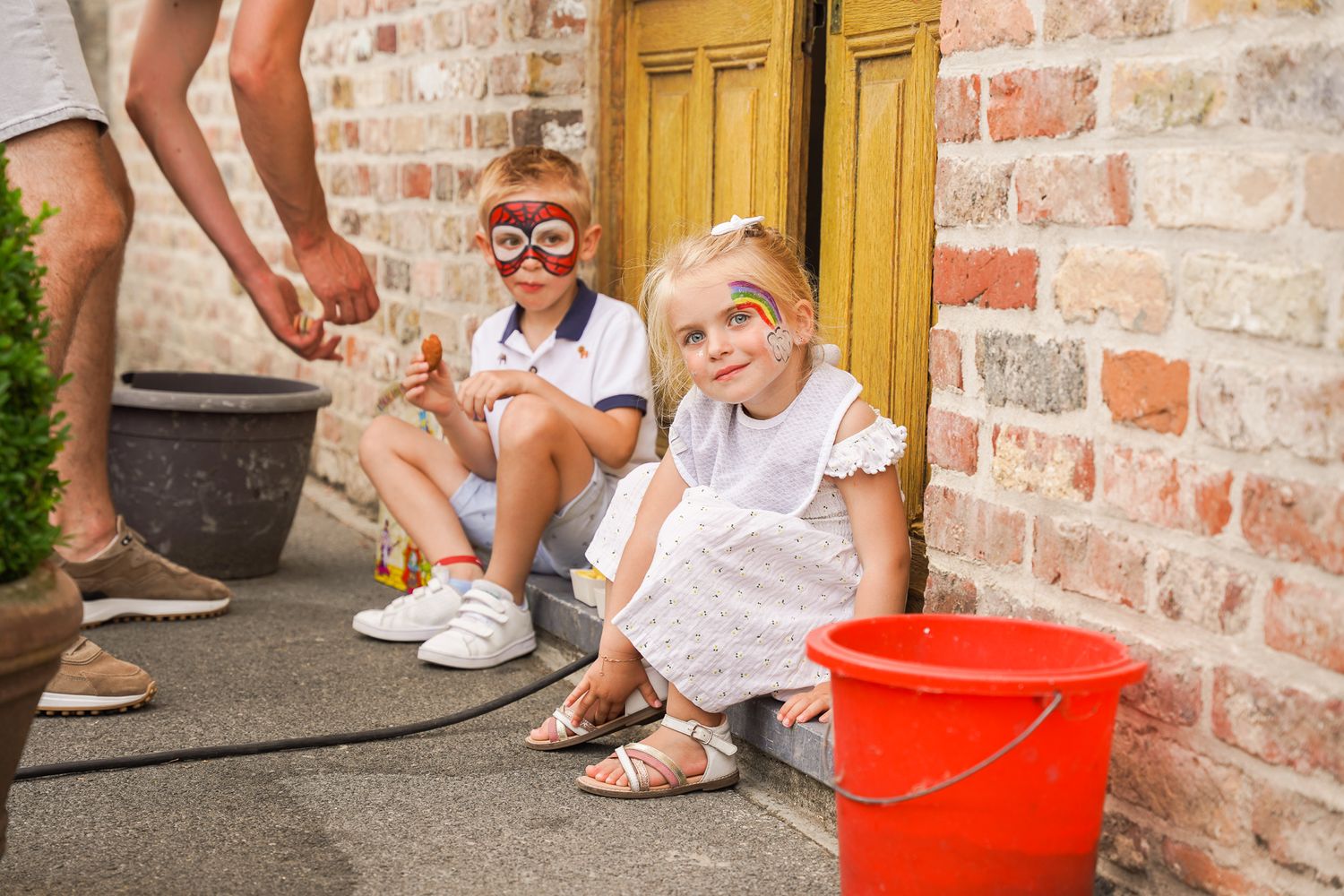 Children sitting on steps with summer outfits and a red bucket against brick wall.