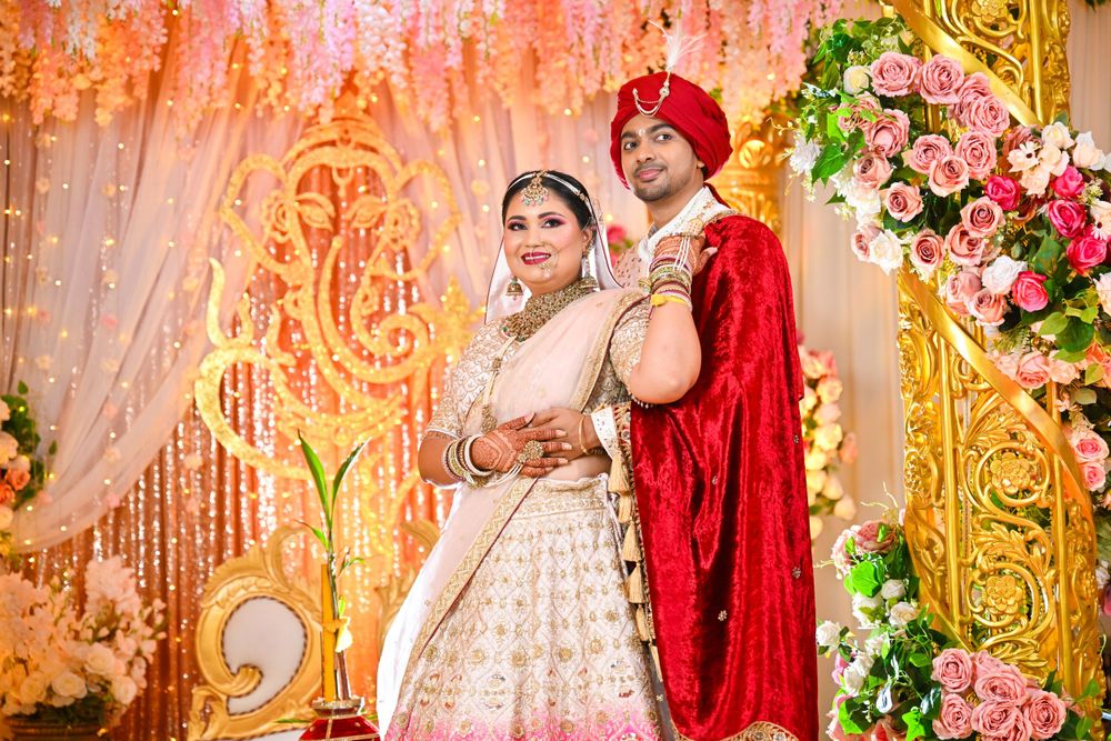 Wedding couple in traditional Indian attire pose against pink floral backdrop.