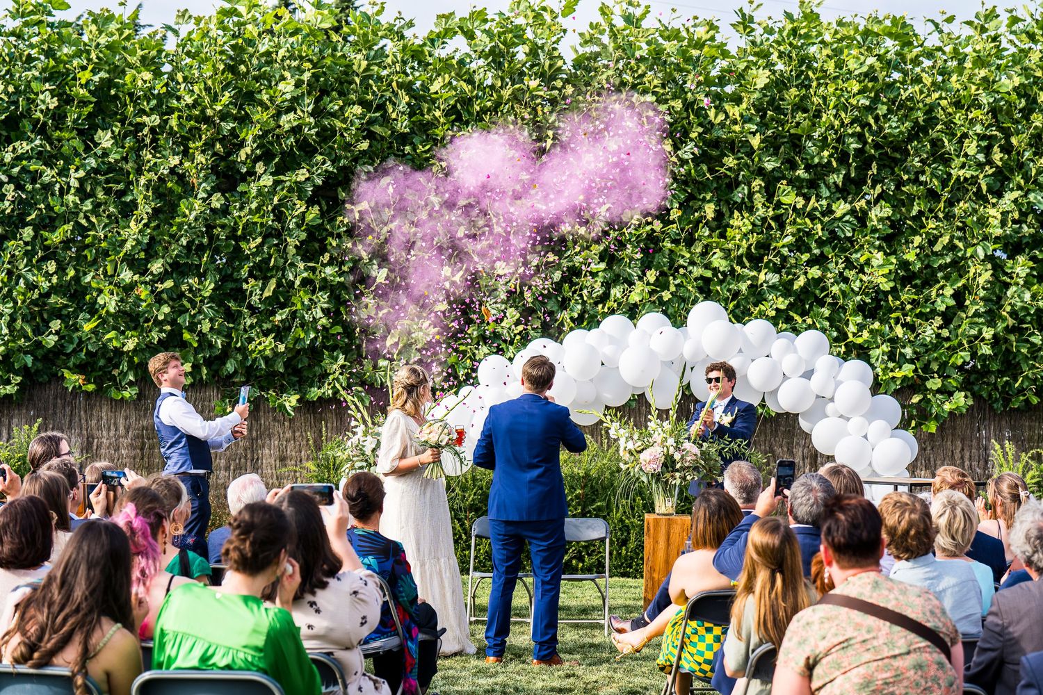 Wedding celebration with pink smoke and white balloon arch against green hedge backdrop.