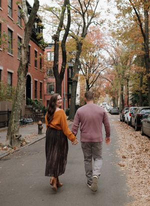 Two people walking together on a tree-lined sidewalk with fallen autumn leaves in a residential neighborhood.