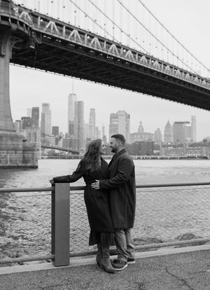 A couple shares a romantic moment along the East River with the Manhattan skyline and bridge visible in the background.
