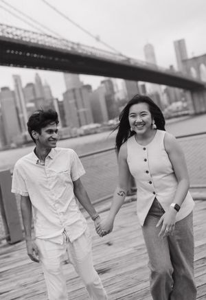 A black and white photo of a couple holding hands and laughing while walking along the waterfront near Brooklyn Bridge.