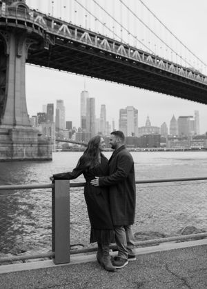 Couple embracing by the East River with Manhattan Bridge and NYC skyline in black and white.