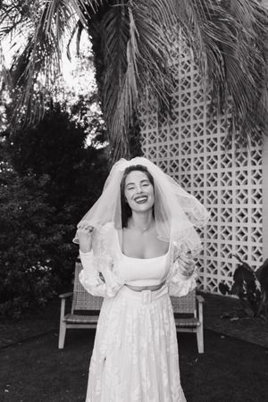 Laughing bride in lace wedding dress and veil poses outdoors near palm tree in black and white photo.