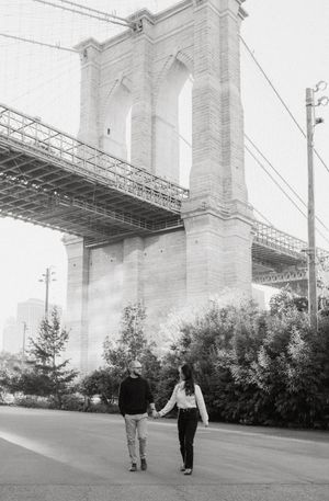 Two people walk beneath the massive concrete pillars of a bridge structure while surrounded by trees and foliage.