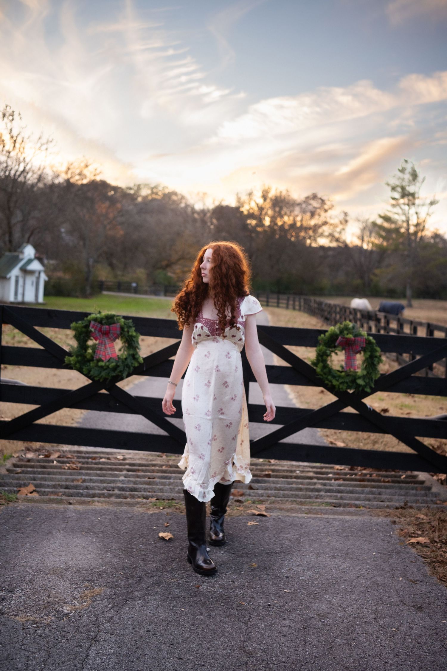 A child in a white dress walks along a rustic wooden fence at sunset in a pastoral countryside setting.