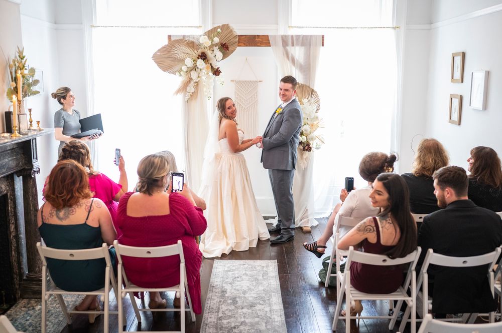 An intimate indoor wedding ceremony in a bright white room with guests seated in chairs watching the couple at the altar.
