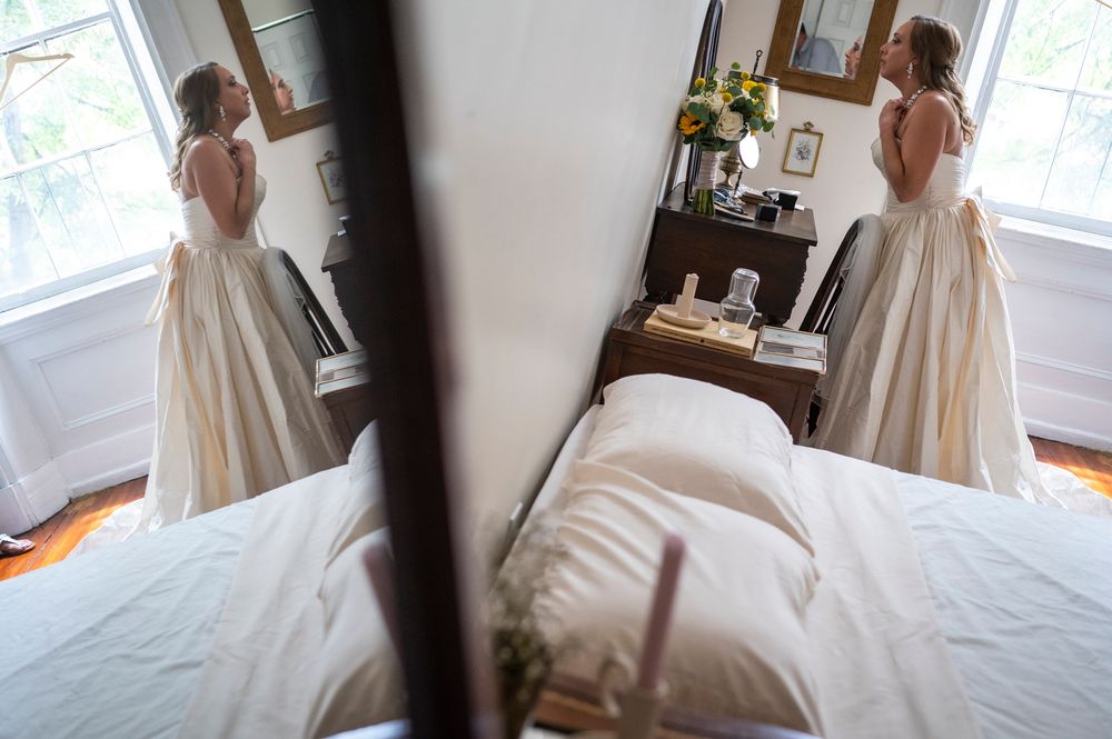 A bride in a flowing white gown stands in a sunlit room looking out the window while holding a bouquet.