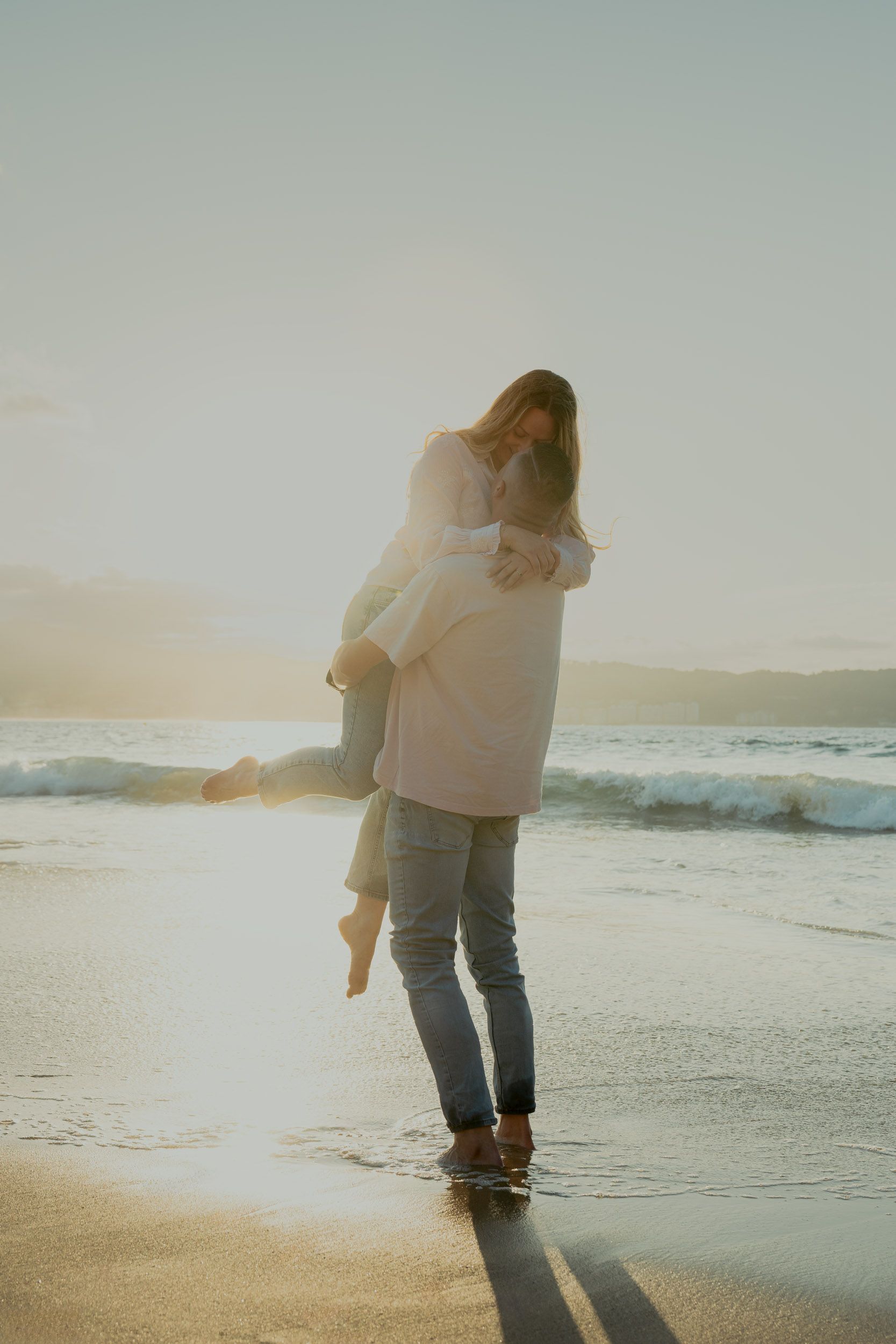 Couple in romantic embrace on beach with golden sunlight streaming through.