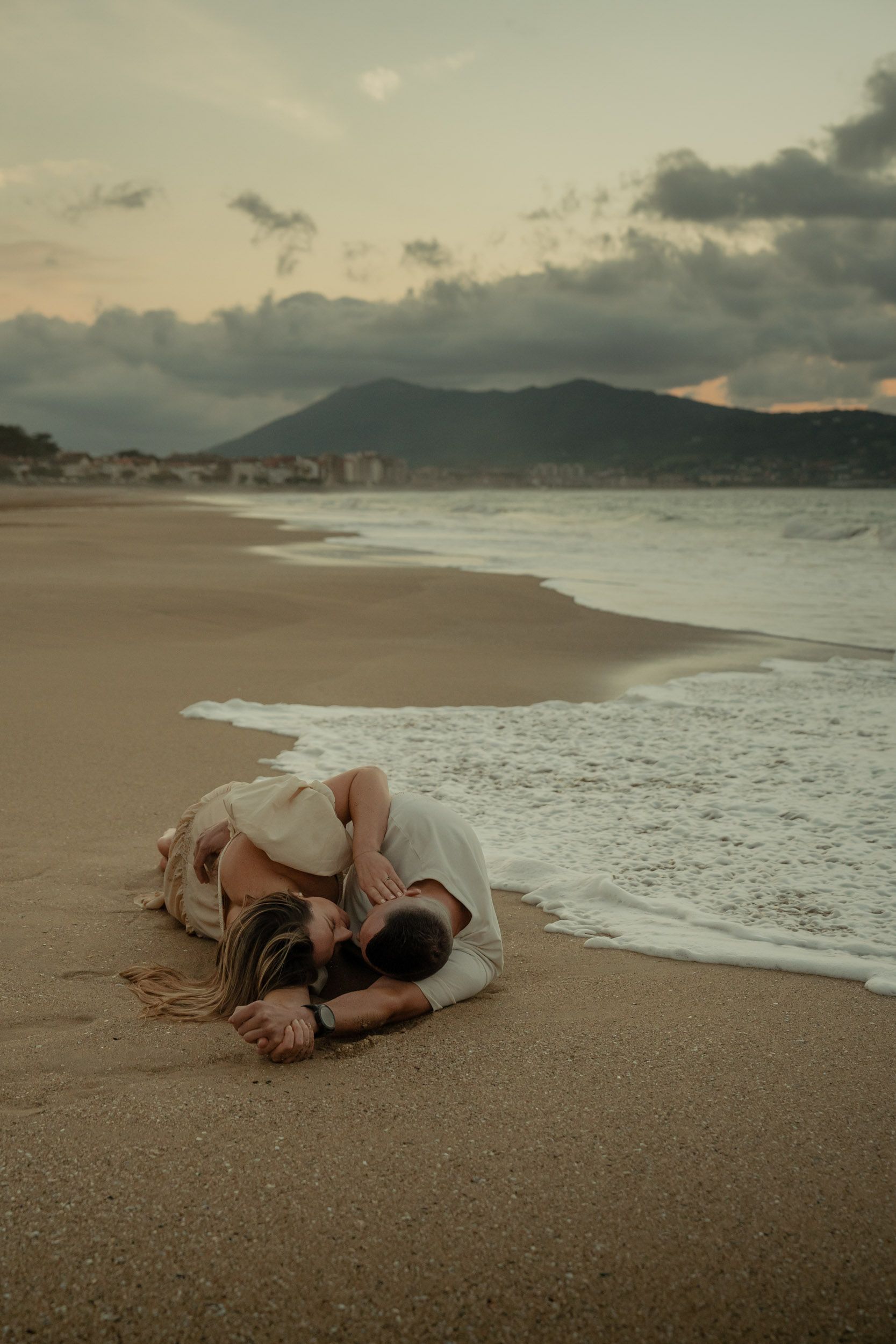 Romantic couple lying on a sandy beach at sunset with waves crashing in the background.