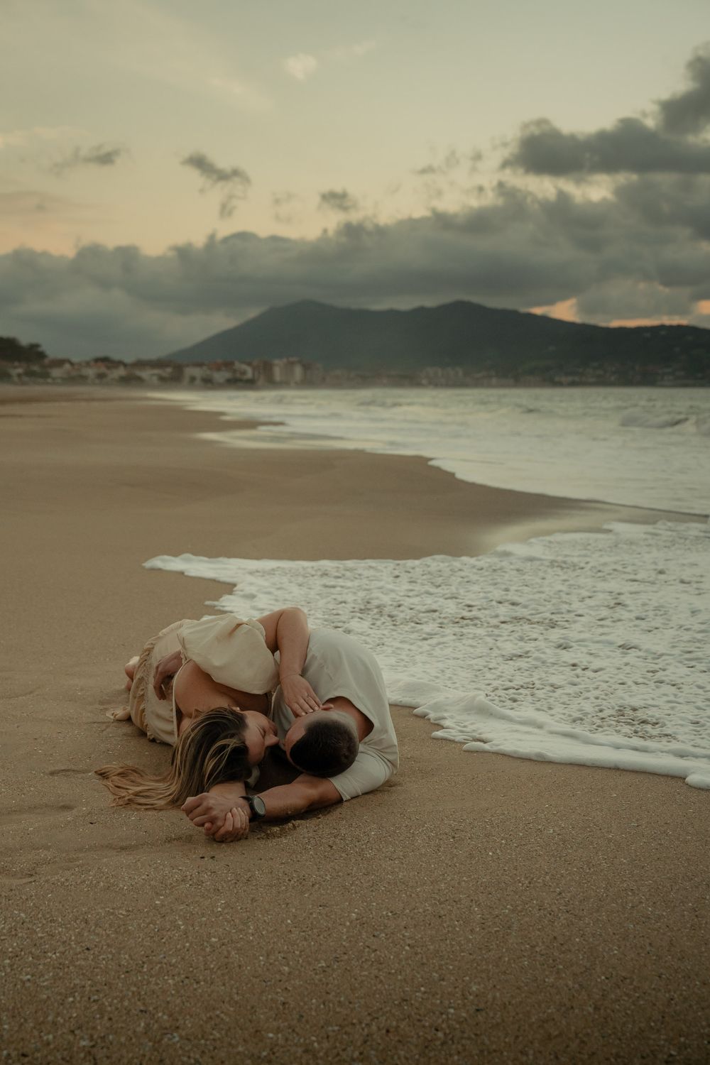 Romantic couple lying on a sandy beach at sunset with waves crashing in the background.
