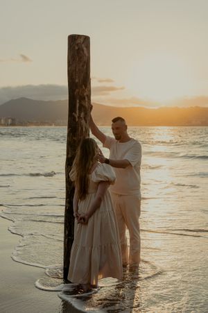 Couple in white clothing embracing by wooden posts in shallow ocean water during golden hour.