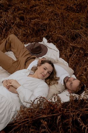Two people lying in white clothing on autumn leaves during a romantic photoshoot.