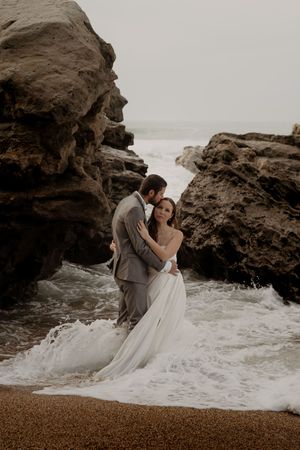 Couple in formal attire embracing near rocky coastal cliffs with crashing waves.
